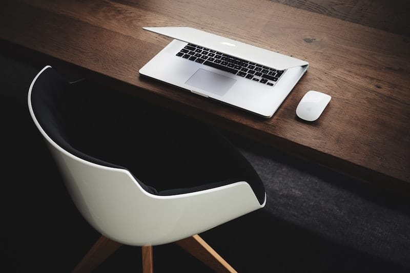 Online Hypnose per Videosprechstunde zeigt Silbernes MacBook mit Apple Maus auf braunem Holztisch mit einem weißen Stuhl und schwarzem Stoff